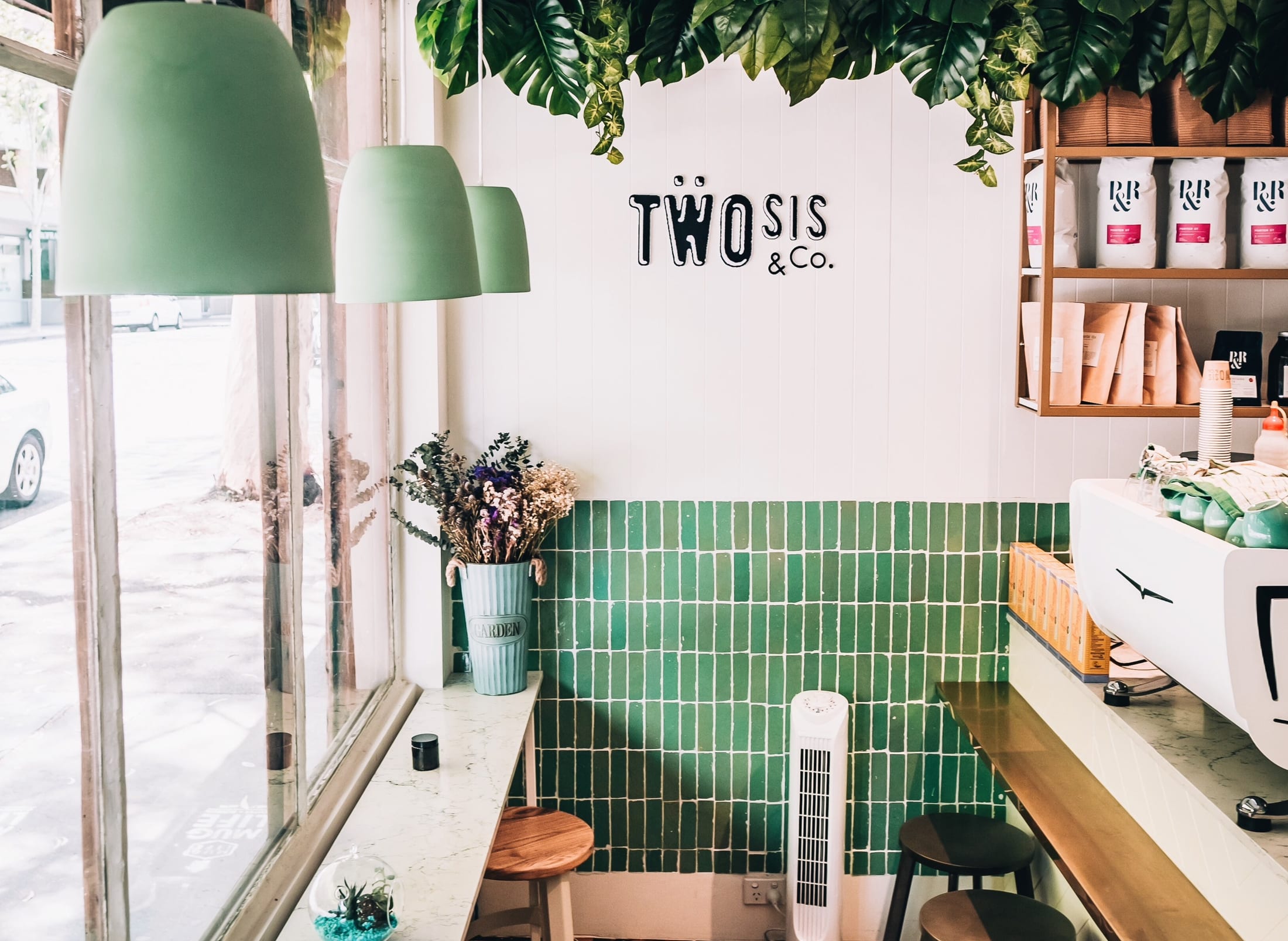 Inside Two Sis and Co: white timber walls, deep green tiles, sage pendant lamps and hanging plants above the coffee bar